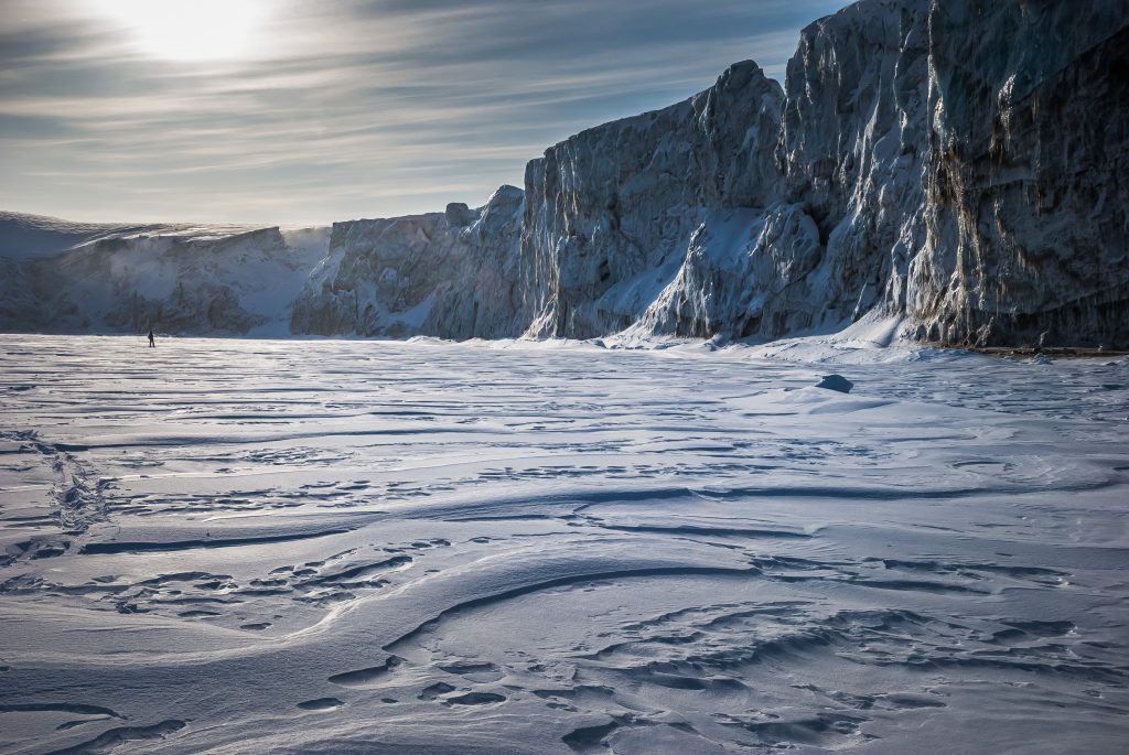 Falaise glacée du Spitzberg dominant une étendue de neige sculptée par le vent, présence humaine minuscule dans l’espace arctique.