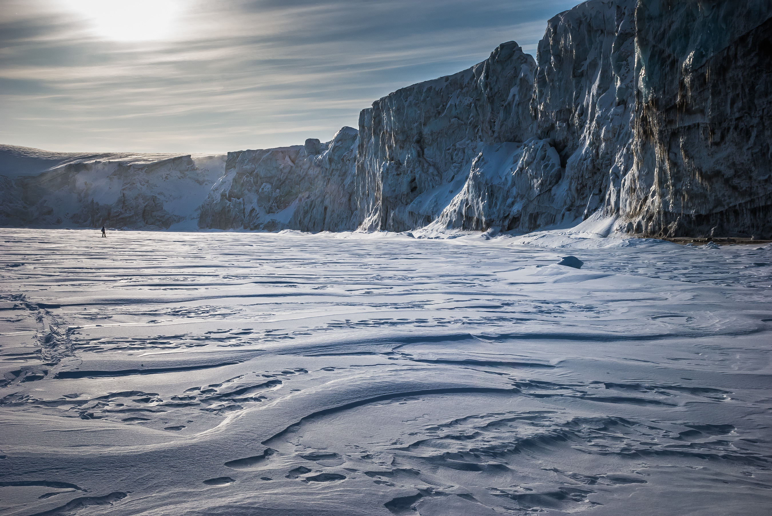Falaise glacée du Spitzberg dominant une étendue de neige sculptée par le vent, présence humaine minuscule dans l’espace arctique.
